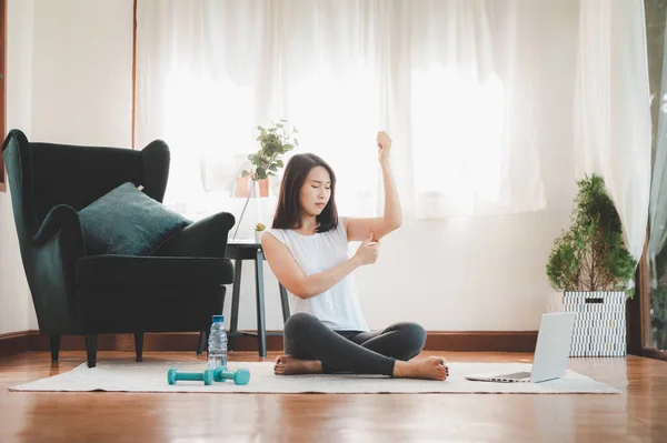 unhappy Asian woman pinching arm tricep fat flabby skin before start workout excercise in living room at home with laptop and dumbbell on the floor