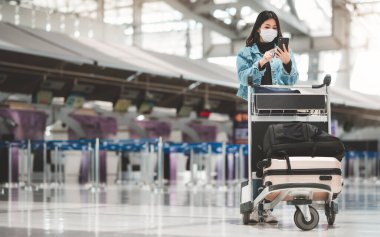 Cep telefonu kullanan Asyalı turist kadın havaalanı terminalinde tek başına bagaj vagonunda duruyor.