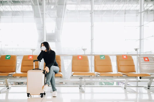 Yüz maskesi takan Asyalı kadın turist, Coronavirus veya Cvid-19 salgını sırasında havaalanı terminalinde bavullarıyla birlikte sosyal mesafeli koltukta oturuyor. Havaalanında yeni normal seyahat