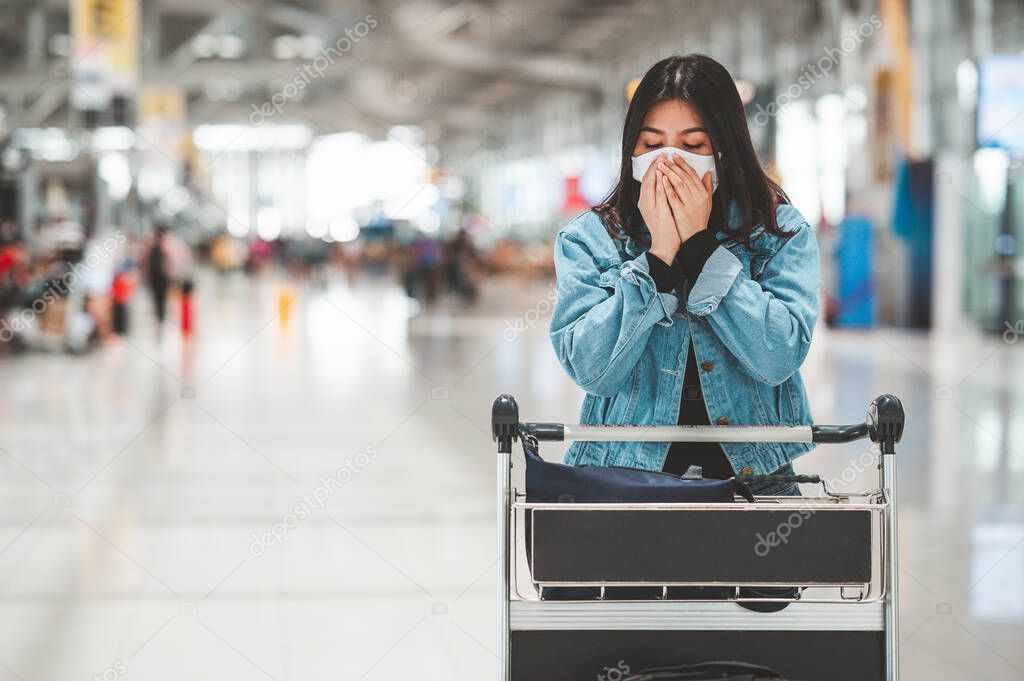 Asian woman tourist wearing protective face mask with luggage trolley sneezing and coughing in airport terminal 