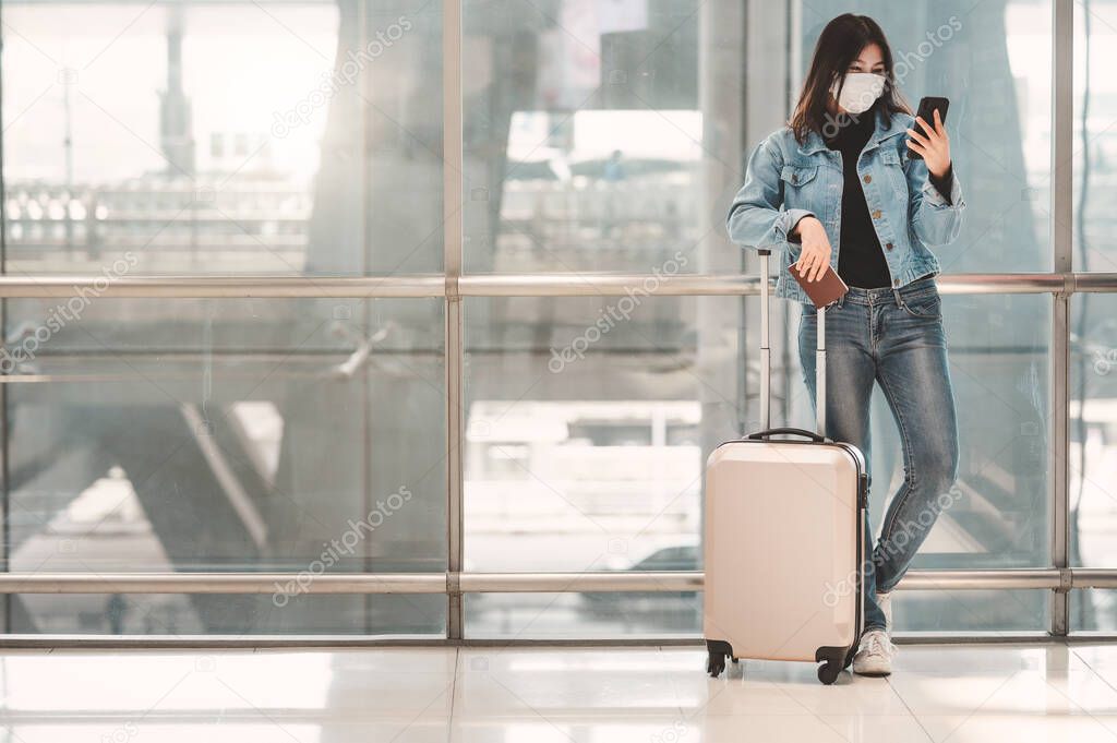 Happy Asian woman tourist with luggage wearing mask protect from coronavirus using smartphone mobile during new normal travel vacation