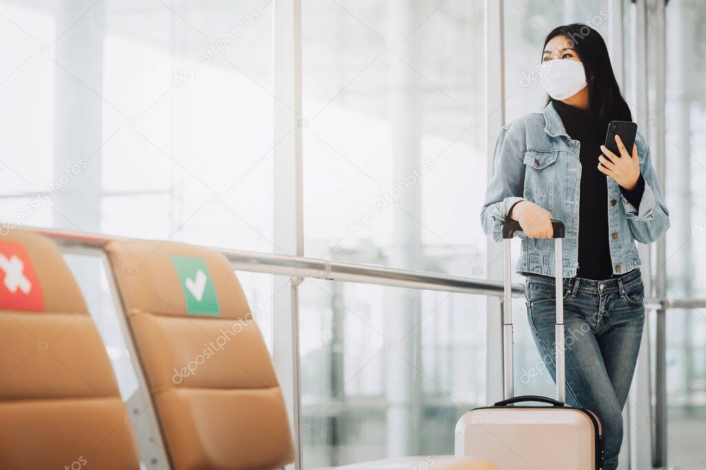 Happy Asian woman traveler wearing mask for protect from coronavirus holding smartphone standing with luggage next to social distancing chair