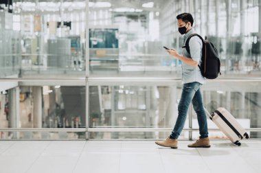 Koronavirüsten korunmak için maske takan Asyalı gezgin elinde akıllı telefon, bavulla yürüyor.