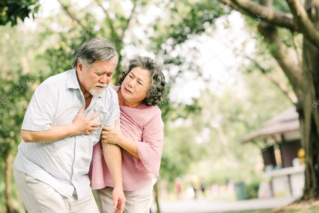 Senior Asian man holding his chest and feeling pain suffering from heart attack while his wife giving support and help outdoor at the park