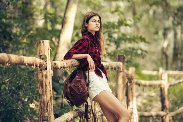 美しい国の少女旅行と赤の市松模様 シャツ ショート パンツ 茶色の革のバックパックを身に着けている木製のフェンス近くに待機 — ストック写真