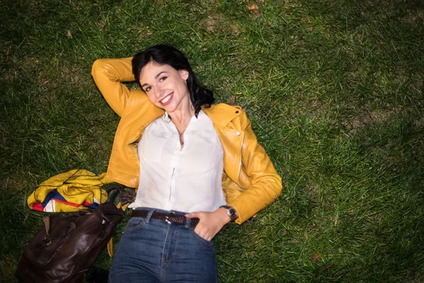幸せな若い女笑顔と夜黄色のジャケットとブルー ジーンズとカラフルな衣装を着て 緑の草の上に横たわる — ストック写真