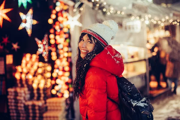 美しい若い女性の笑みを浮かべて楽しむ雪のクリスマス夜市の身に着けている帽子と赤いジャケット フェア冬時間 — ストック写真