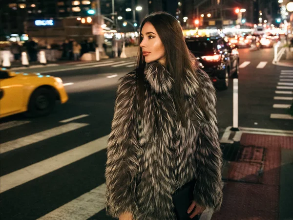 夜の街の偽の毛皮のジャケットと側にいる保持袋を身に着けている通りの上を歩いてゴージャスなエレガントな女性 — ストック写真
