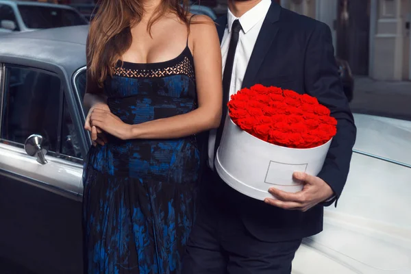 ロマンチックなカップルの日付 男性と女性の身に着けているバラの花に合わせて 都市で高級車近くに立ってのドレス — ストック写真