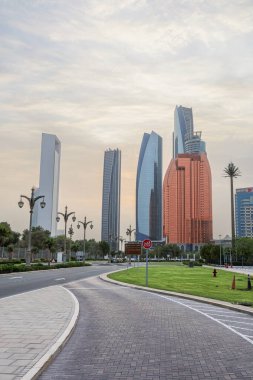 Abu Dabi, BAE 16 Ağustos 2025: Gökdelenler Birleşik Arap Emirlikleri 'nin başkentinde gün batımında fıskiyelerle dolu palmiye dolu bir parka bakıyor.