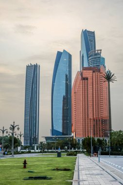 Abu Dabi, BAE 16 Ağustos 2025: Gökdelenler Birleşik Arap Emirlikleri 'nin başkentinde gün batımında fıskiyelerle dolu palmiye dolu bir parka bakıyor.