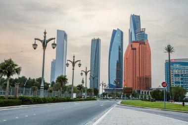 Abu Dabi, BAE 16 Ağustos 2025: Gökdelenler Birleşik Arap Emirlikleri 'nin başkentinde gün batımında fıskiyelerle dolu palmiye dolu bir parka bakıyor.