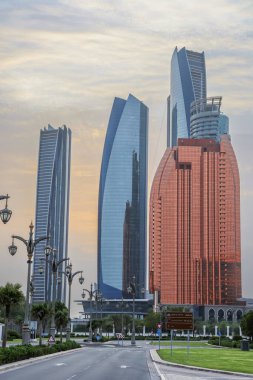 Abu Dabi, BAE 16 Ağustos 2025: Gökdelenler Birleşik Arap Emirlikleri 'nin başkentinde gün batımında fıskiyelerle dolu palmiye dolu bir parka bakıyor.