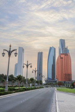 Abu Dabi, BAE 16 Ağustos 2025: Gökdelenler Birleşik Arap Emirlikleri 'nin başkentinde gün batımında fıskiyelerle dolu palmiye dolu bir parka bakıyor.