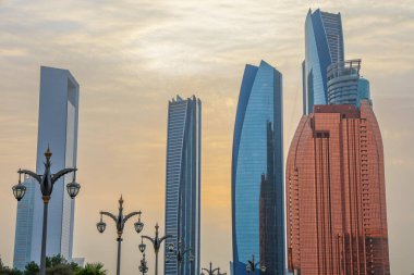 Abu Dabi, BAE 16 Ağustos 2025: Gökdelenler Birleşik Arap Emirlikleri 'nin başkentinde gün batımında fıskiyelerle dolu palmiye dolu bir parka bakıyor.