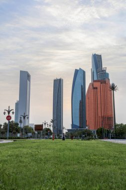 Abu Dabi, BAE 16 Ağustos 2025: Gökdelenler Birleşik Arap Emirlikleri 'nin başkentinde gün batımında fıskiyelerle dolu palmiye dolu bir parka bakıyor.