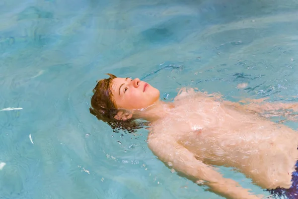 屋内プールで泳いでいると 背中に横たわって 赤い髪 Enyoing を持つ少年 — ストック写真