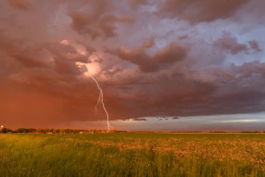 A large dust storm blows across a field and small town at sunset, casting a vibrant deep orange hue across the horizon and topped off with a large lightning bolt clipart