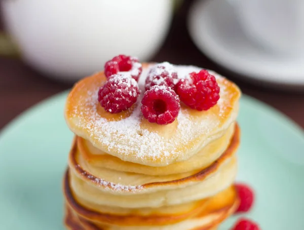 Stack of baked homemade pancakes with raspberries, sprinkled with powdered sugar and tea utensils in the background, selective focus — Stock Photo, Image