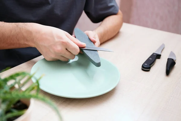 Sharpening the knife with whetstone. Kitchen appliance care. Close-up strong male hands sharpen a kitchen metal knife with a grindstone. Home household knife sharpening.