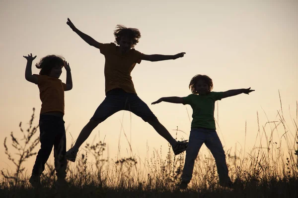 Dzieci bawiące się skoki na lato zachód słońca łąka sylwetki — Zdjęcie stockowe