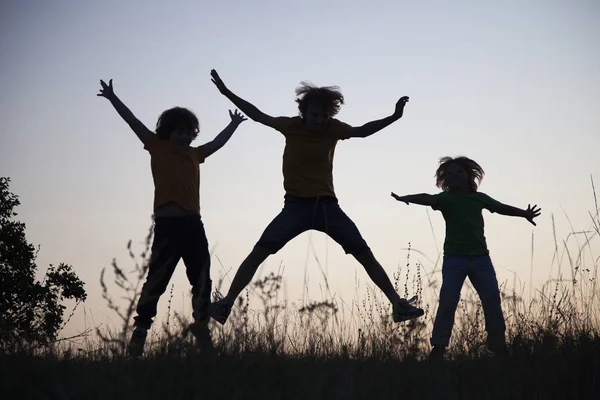 Niños jugando saltar en verano puesta del sol pradera silueta —  Fotos de Stock