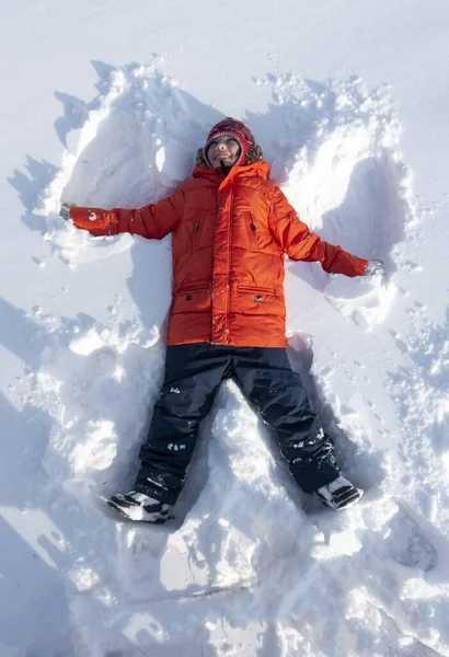 Menino feliz deitado na neve e fazendo figura de anjo da neve com as mãos — Fotografia de Stock