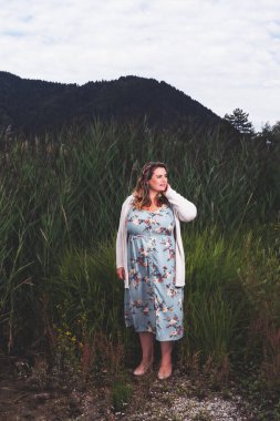 Büyük göğüslü kıvrımlı bir kadın, göl kenarında yaz elbisesi içinde duruyor..