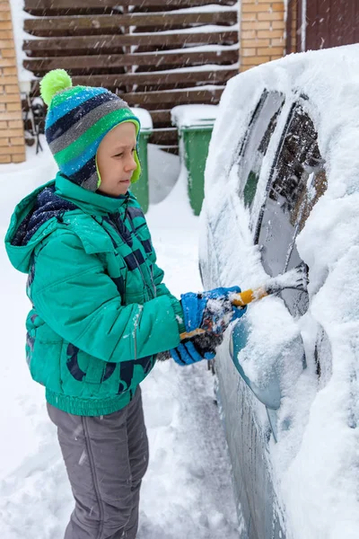 男孩站在车边拿着刷子 孩子们从雪中清洗汽车 暴风雨过后把汽车上的雪刷干净 — 图库照片
