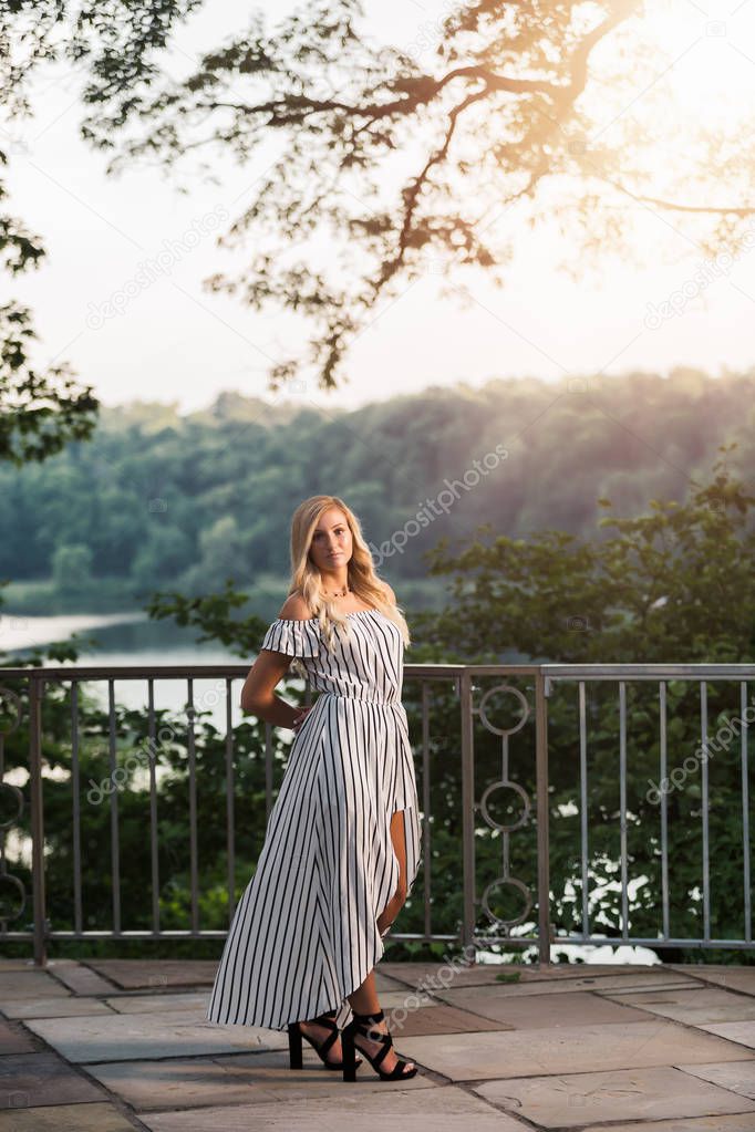 One attractive blonde hair caucasian high school senior posing for senior pictures. Female Teenager portrait standing outdoors at park in summer.