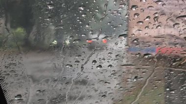 Yağmur damlalarıyla kaplı bir arabanın yan aynasına yakın çekim, ıslak bir yolu ve farları açık beyaz bir aracı yansıtıyor. Bulanık arkaplan ve su damlacıkları yağmurlu hava ve sürüş koşullarını anımsatıyor.