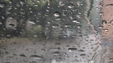 Yağmur damlalarıyla kaplı bir arabanın yan aynasına yakın çekim, ıslak bir yolu ve farları açık beyaz bir aracı yansıtıyor. Bulanık arkaplan ve su damlacıkları yağmurlu hava ve sürüş koşullarını anımsatıyor.