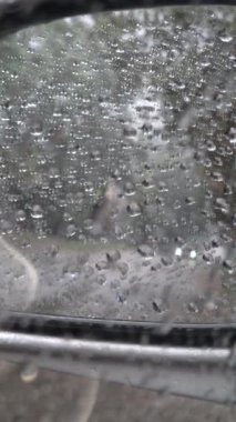 Yağmur damlalarıyla kaplı bir arabanın yan aynasına yakın çekim, ıslak bir yolu ve farları açık beyaz bir aracı yansıtıyor. Bulanık arkaplan ve su damlacıkları yağmurlu hava ve sürüş koşullarını anımsatıyor.