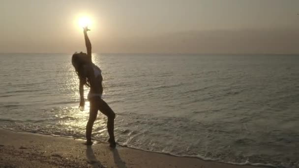 Σιλουέτα της νεαρής γυναίκας με μακριά μαλλιά χορεύει στο ανάχωμα πάνω από τον ωκεανό ή τη θάλασσα κατά την Ανατολή ή το ηλιοβασίλεμα.. Αργή κίνηση. — Αρχείο Βίντεο