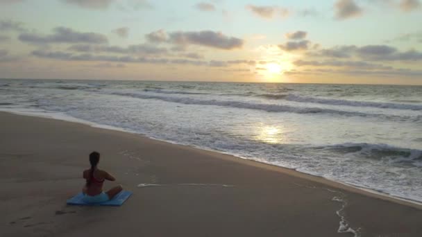 Hermosa joven haciendo yoga sentado en la playa mientras observa el atardecer . — Vídeo de stock