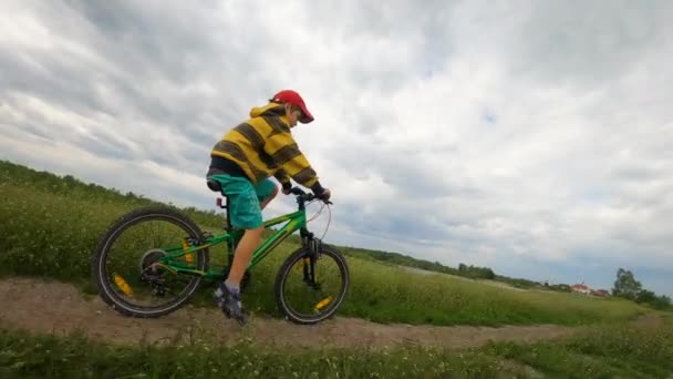 Un niño monta en bicicleta en un camino de tierra a lo largo del río. Clima nublado, nubes de tormenta . — Vídeos de Stock