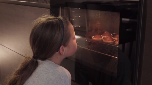 Девушка смотрит на духовку на кухне и ждет печенье — стоковое видео