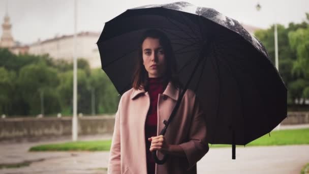 Молодая серьезная женщина, стоящая под зонтиком под дождем, грустная погода в городе — стоковое видео