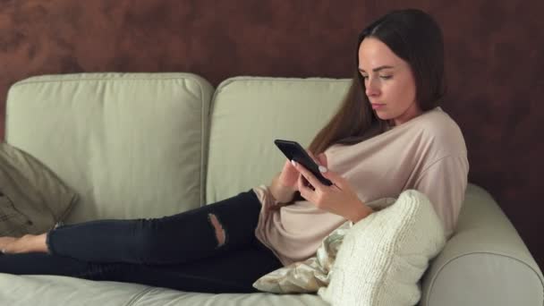 Ung kvinna med smartphone som sitter på soffan hemma — Stockvideo