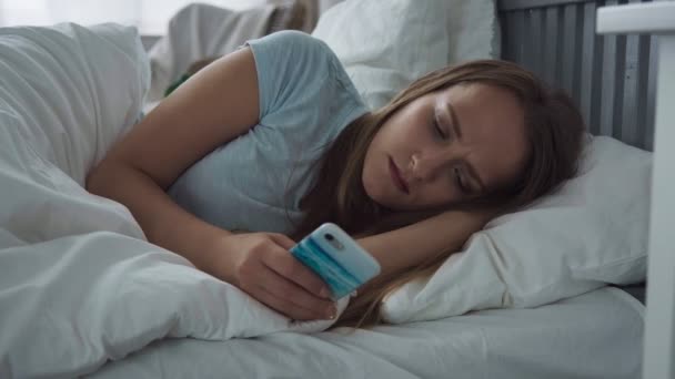 Уставшая женщина с грустными глазами с помощью смартфона на кровати дома — стоковое видео