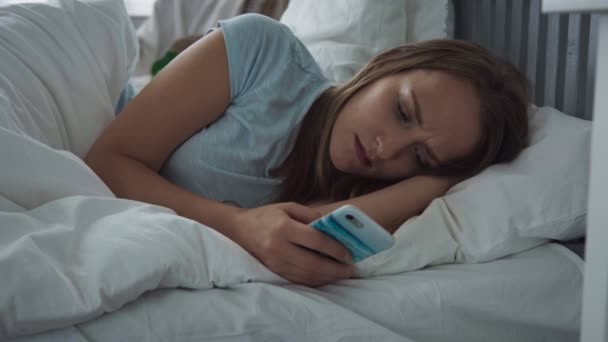 Λυπημένη γυναίκα ξαπλωμένη στο κρεβάτι, χρησιμοποιώντας το smartphone και προσπαθεί να κοιμηθείτε — Αρχείο Βίντεο