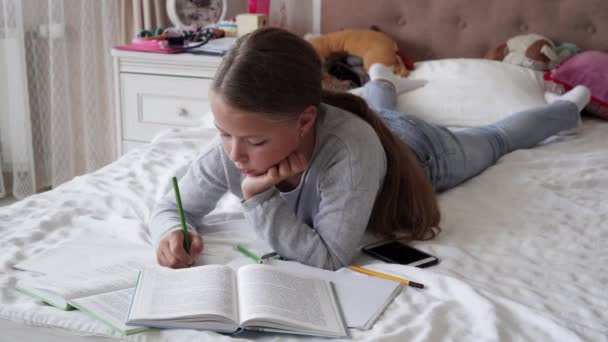 Маленька школярка лежить на ліжку і робить домашнє завдання — стокове відео