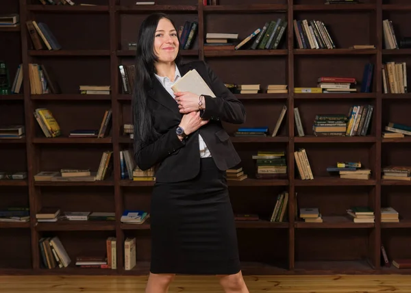 Atractiva mujer sosteniendo libro y sonriendo cerca de la estantería — Foto de Stock