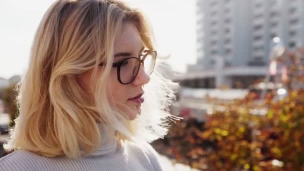 Όμορφη νεαρή ξανθιά γυναίκα με τα γυαλιά μακριά κοιτάζοντας ηλιόλουστη φθινοπωρινή πόλη — Αρχείο Βίντεο