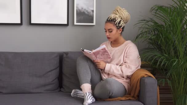Молодая женщина пишет в блокноте, сидя на удобном диване дома — стоковое видео