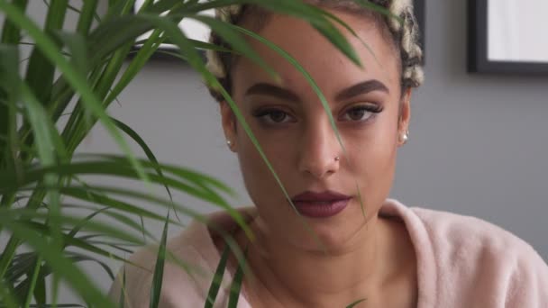 Happy young woman with beautiful eyes smiling through leaves of plant in room — Stock Video