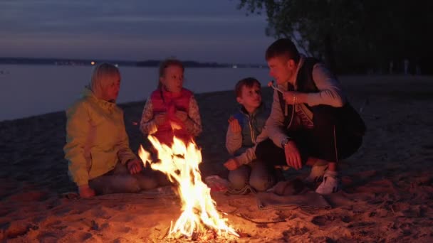 Familia feliz comiendo y hablando por el fuego durante el picnic en la playa de la noche — Vídeo de stock