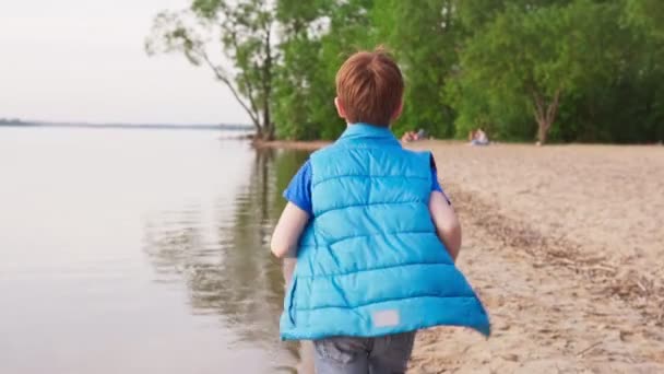 Мальчик бегает по песчаному пляжу вдоль береговой линии в летний день — стоковое видео