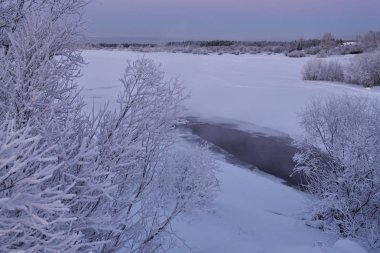 Doğa, kış soğuk akşam güneş battıktan sonra nehir kıyısında. Ağaçlar nem dondan kalın bir tabaka ile. Nehir henüz tamamen donmuş değil.