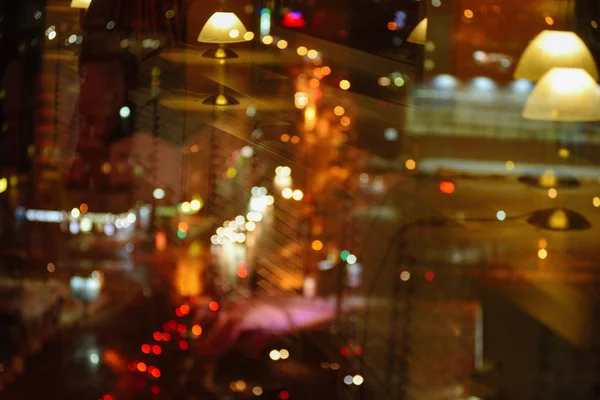 View of the night streets of the city from the window of a tall building with a reflection in the glass window of table lamps.